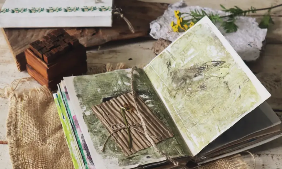 Otwarty notatnik do art journalingu z zielonymi, abstrakcyjnymi wzorami. Na jednej ze stron znajduje się suszony kwiat na pogniecionej tekturze.