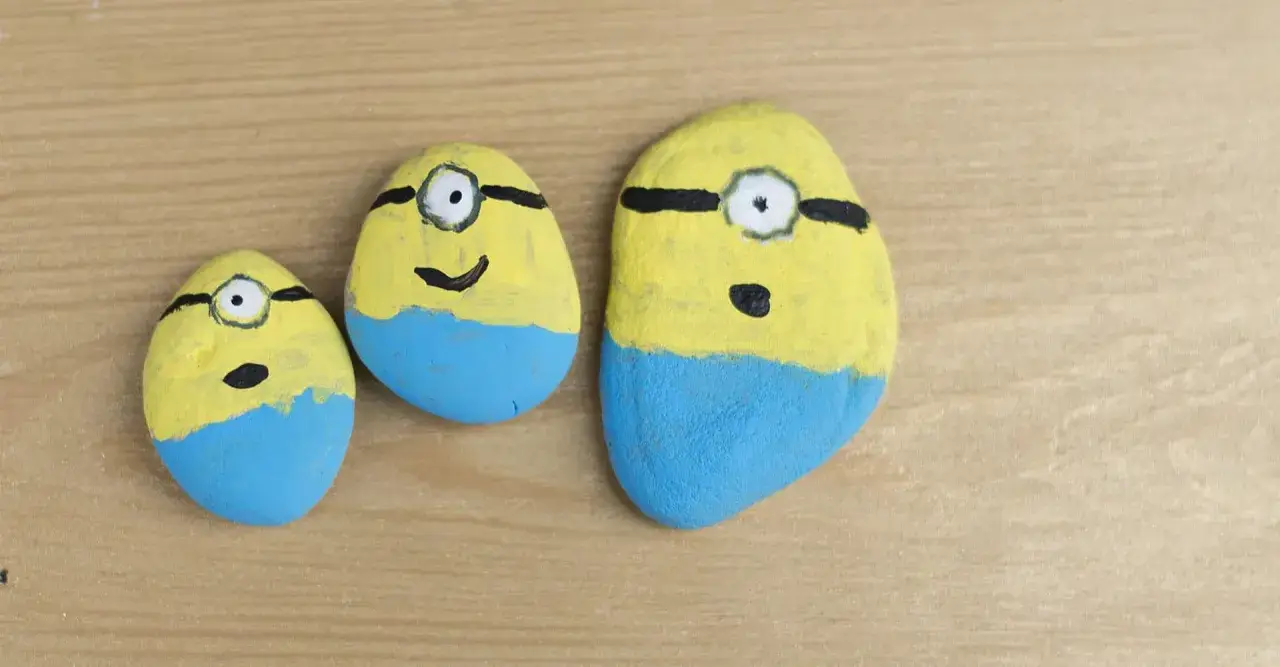 Urocze malowanie kamieni w kształcie Minionków. Trzy kamienie, każdy z żółtą górą, niebieskimi spodenkami i jednym okiem.