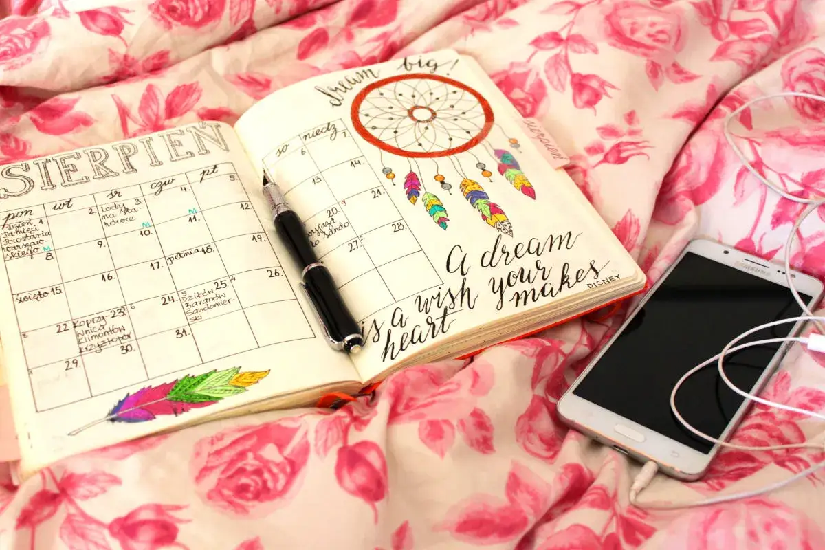 Bullet journal co to? Otwarty notatnik z kalendarzem na sierpień, pióro, telefon i łapacz snów.