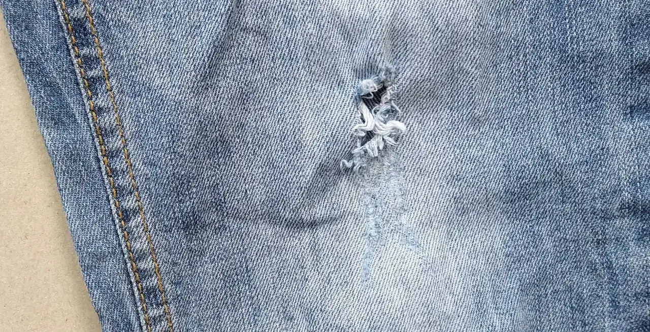 Zbliżenie na jasne jeansy z widoczną dziurą, kt&oacute;ra czeka na to, jak zaszyć dziurę w spodniach. Widoczny szew boczny.