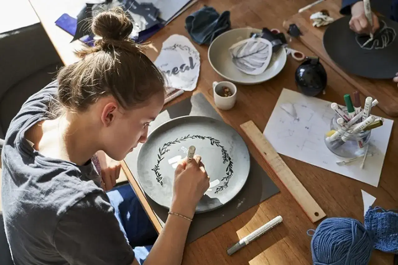 Dziewczyna maluje wzór na talerzu, jakby tworzyła własny zimna porcelana przepis. Na stole materiały do rękodzieła.