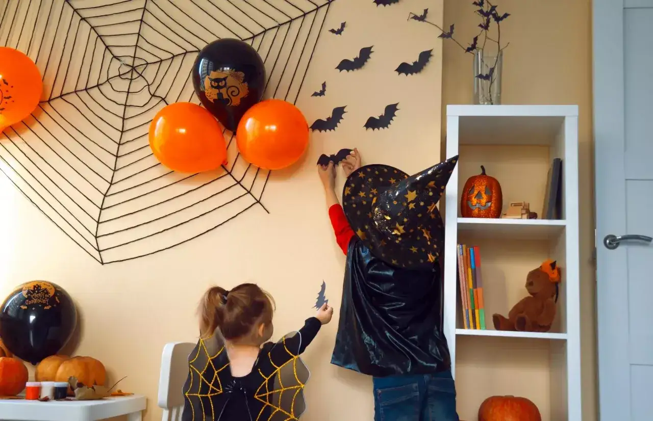 Dzieci dekorują pokój na Halloween. Na ścianie wisi wielka pajęczyna, balony i nietoperze.